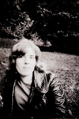 Portrait of a person with short, wavy hair resting outdoors on grass, framed by dense foliage. Mid-century style clothing sug...
