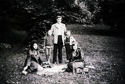 Family portrait on a grassy outdoor setting, likely 1960s–1970s. Five individuals pose: two adults (standing) and three child...