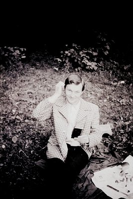 Black-and-white candid of a person sitting on forest floor, holding a glass of wine and a book. Mid-20th century outdoor port...