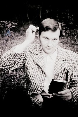Black-and-white portrait of a man in a plaid shirt, seated outdoors among foliage. He holds a small object (likely a book or ...