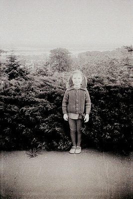 Black-and-white portrait of a young girl standing on a gravel path beside dense evergreen foliage, likely a coastal or park s...
