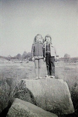 Two children pose on a rocky outcrop in a rural landscape, mid-20th century. Girl in striped sweater and boy in jacket with h...