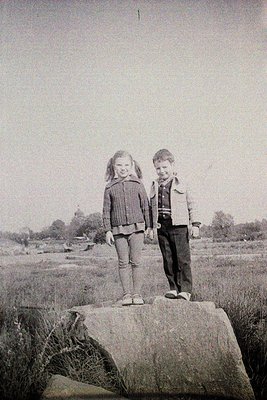 Two children pose on a rocky outcrop in mid-20th century clothing—girl in pleated skirt, striped sweater, and cardigan; boy i...