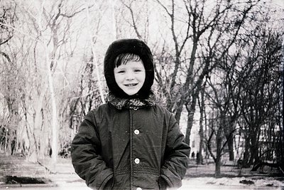Mid-century black-and-white portrait of a smiling child in winter attire—fur-lined hood and buttoned coat—standing on a snow-...