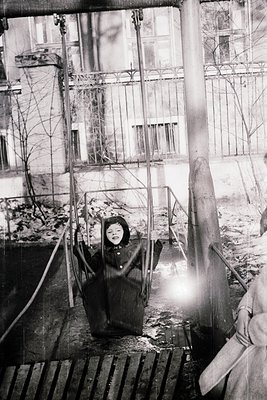 Child in heavy winter coat stands on elevated wooden platform beside rusted metal railings, framed by a multi-story building ...