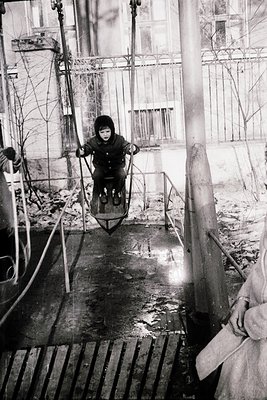 Mid-20th century child riding a hand-pulled sled on a wet, slatted wooden platform, likely a dock or pier. Industrial pipes a...
