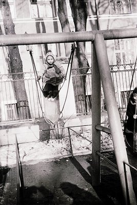 Child mid-swing on a rope swing in an urban courtyard, framed by wrought-iron railings. Snow covers ground and nearby benches...