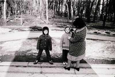 Black-and-white snapshot of three children in winter attire on a paved path, flanked by bare trees. The adult holds a camera,...