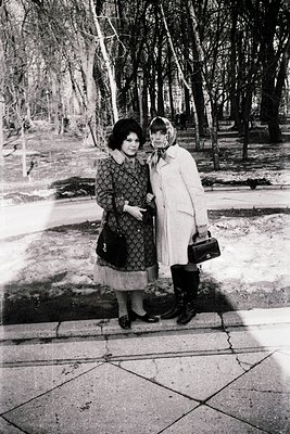 Two women pose on a paved pathway in a park, likely mid-20th century (1960s–1970s). The woman on the left wears a patterned d...