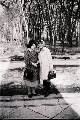 Two women pose closely in a park during winter, 1960s. One wears a patterned dress with a collared coat, the other a white co...