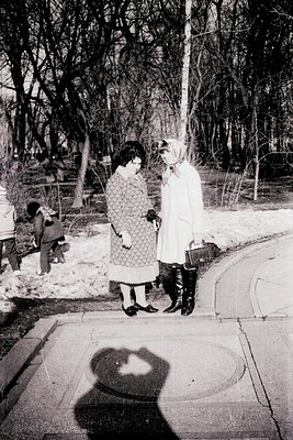 Two women in winter attire converse outdoors, likely mid-1960s. One wears a patterned dress with knee-high boots; the other, ...