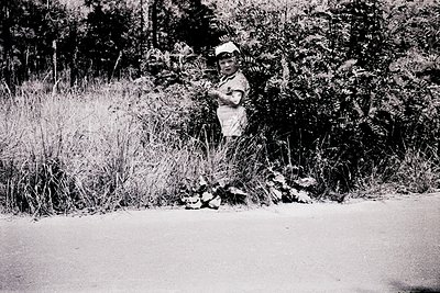 Child in vintage outdoor setting, mid-20th century. Black-and-white photo captures a young boy in a white shirt and cap, hold...