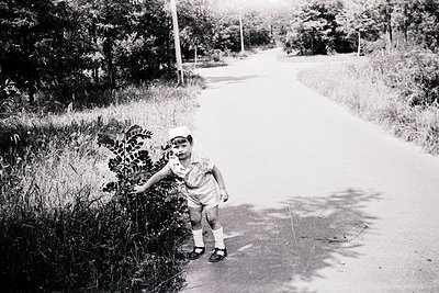 Black-and-white snapshot of a young child in mid-1960s rural setting, standing on paved roadside. Child wears short-sleeved s...