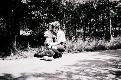 A woman kneels beside a child in a wooded area, both dressed in casual 1970s attire—she wears a light blouse, dark pants, and...