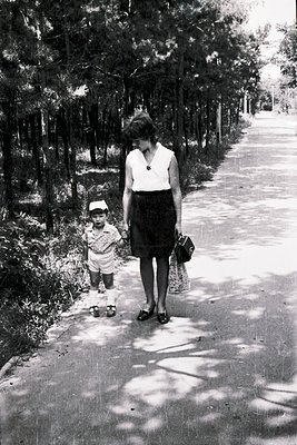 A woman in a knee-length skirt and blouse, holding a child’s hand, walks a tree-lined path in mid-20th century attire. The ch...