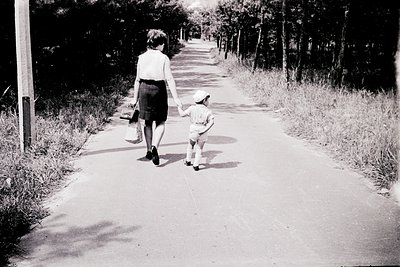 Black-and-white street scene: adult and child walking hand-in-hand on a paved path lined with trees and wild grass. Mid-20th ...