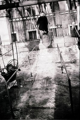 Mid-20th-century urban construction site with a lone worker mid-air on a metal scaffold, performing maintenance or repair. In...