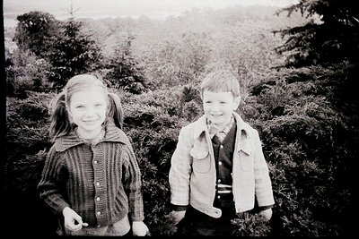 Vintage black-and-white portrait of two children posing outdoors in a forested area, likely mid-20th century. Girl in knitted...