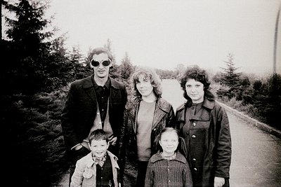 Black-and-white portrait of a family of five posing outdoors on a paved path surrounded by trees. The man wears sunglasses an...