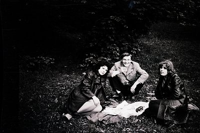 Three individuals pose casually on forest floor, mid-1970s. Central figure wears plaid shirt and dark trousers; left and righ...
