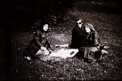 Three individuals seated on forest floor, mid-20th century. Left: woman in long coat, dark hair. Center: man in suit, sunglas...