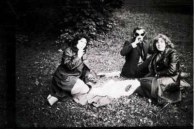 Three individuals pose playfully outdoors in 1960s-70s fashion—one in sunglasses, another with hands over mouth, all seated o...