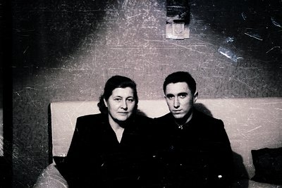 Vintage black-and-white portrait of two individuals seated closely in an indoor setting, likely mid-20th century. The woman w...