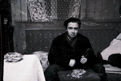 Black-and-white interior shot of a man seated on a wooden stool, holding a hammer and small metal object, likely a nail or sc...