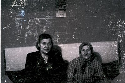 Two women pose indoors on a worn, cracked sofa against a textured wall. The woman on the left wears a dark coat; the right we...