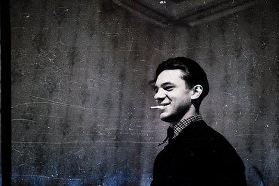 Portrait of a man in mid-20th century attire—dark blazer, checkered shirt, and cigarette holder—set against a textured, aged ...