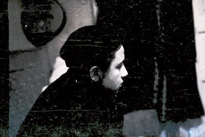 Vintage black-and-white portrait of a man in profile, seated against a textured wall with graffiti-like markings. His attire ...