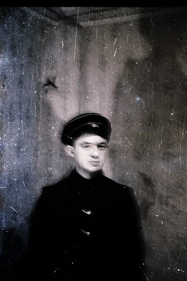Vintage sepia-toned portrait of a young man in a peaked cap and dark coat, likely mid-20th century. The aged, textured wall a...