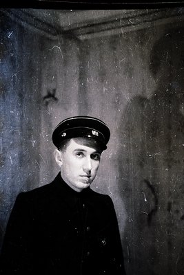 Black-and-white portrait of a young man in a peaked cap and dark formal attire, likely mid-20th century. The aged, textured b...