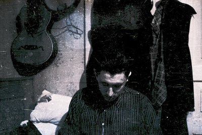 Vintage black-and-white photo of a person seated indoors, holding a guitar with a large, stylized face painted on its body. T...