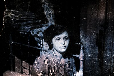 Vintage sepia-toned portrait of a woman in a patterned blouse, leaning against a rustic wooden structure. Evidence of age and...