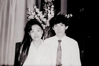 Black-and-white portrait of two individuals in formal attire, likely from the 1970s–1980s. The man wears a white dress shirt ...