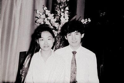 Two individuals pose formally in a black-and-white photo, likely from the 1970s–1980s. The woman wears a loose blouse with a ...