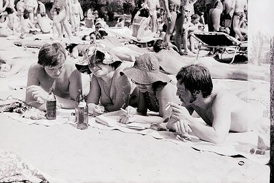 Vintage beach scene with four adults seated on a blanket, enjoying drinks and snacks. 1970s summer attire—women in sun hats a...