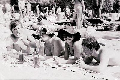 Black-and-white beach scene from the 1970s, featuring four relaxed individuals seated at a picnic table. Sunbathing crowd and...