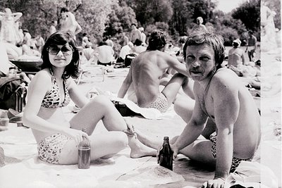 Black-and-white beach scene from the 1970s, featuring relaxed summer attire—women in floral swimsuits, men in patterned swim ...
