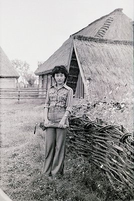 Young woman in 1970s-style wide-legged trousers and button-up blouse stands beside a thatched-roof hut, holding a bundle of s...