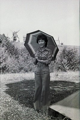 Vintage black-and-white portrait of a woman in 1970s-style wide-leg trousers and plaid shirt, holding a striped umbrella outd...