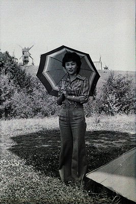 1970s portrait of woman in wide-leg trousers and plaid shirt, holding a striped umbrella in a rural Dutch landscape with wind...
