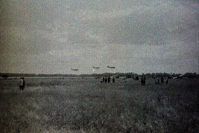 Vintage sepia-toned aerial display over open grassland with four biplanes in formation. Crowd of onlookers gathered in foregr...