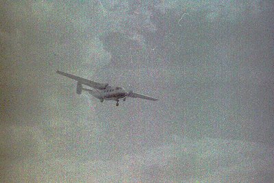 Vintage black-and-white photo of a propeller-driven aircraft in mid-flight, likely a twin-engine turboprop. The aircraft’s hi...