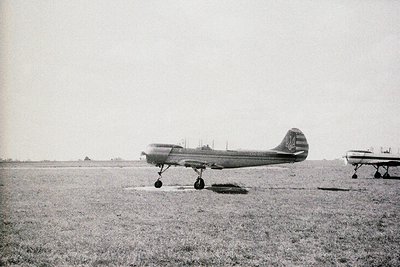 Vintage biplane on grass airfield, mid-20th century. High-wing monoplane with streamlined fuselage, visible propeller, and ta...