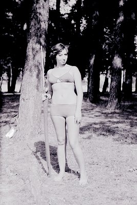 Vintage black-and-white portrait of a woman in a one-piece swimsuit, posing outdoors near a tree trunk. The sleek, form-fitti...