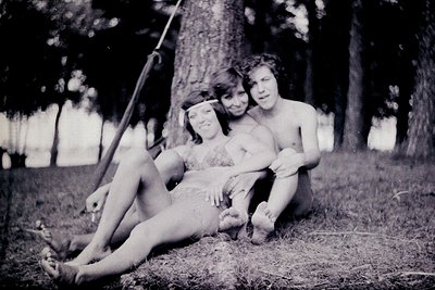 Vintage black-and-white photo of three young people posing on a swing in a wooded area, mid-20th century. The swing hangs fro...