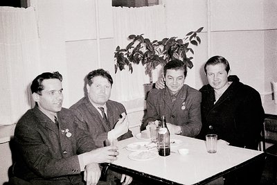 Four men in formal 1950s–60s attire pose indoors around a table with food/drinks. Soviet-style medals and badges visible on l...