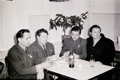 Four men in mid-20th-century formalwear—dark suits, ties, and medals—sit at a simple rectangular table in an indoor setting, ...